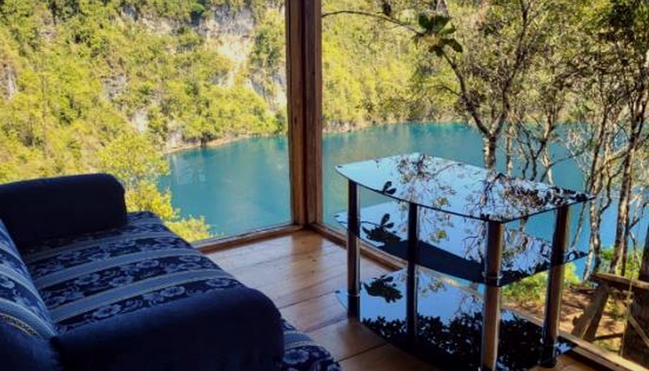 Interior de cabaña de cristal con ventanales y vista al lago. Usar como hero de la sección.