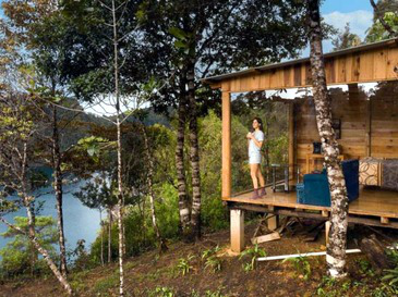 Exterior de cabaña de cristal con terraza y vista al lago.